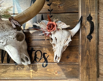 May include: A wall-mounted wooden sign featuring two white animal skulls adorned with dried flowers, lace, and a rust-colored rose. The dark brown horizontal wood planks display black text including the word 'MATOS' and 'EST 2012' with a black metal handle.