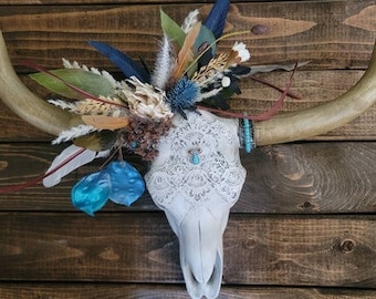 May include: A decorative cow skull with long horns, adorned with a floral arrangement of feathers, dried flowers, and blue accents. The skull is painted white and overlaid with a lace pattern, and a turquoise stone is set in the center.