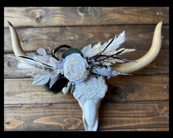 May include: White faux taxidermy bull skull with a white lace overlay and a floral arrangement of white and cream flowers, dried grasses, and black feathers.