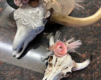 May include: Two white cow skulls decorated with lace and pink flowers. The skulls are on a brown surface.