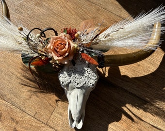 May include: A decorative faux cow skull with long horns, adorned with a floral arrangement. The arrangement includes a large peach rose, dried grasses, and other natural elements. A white lace detail is attached to the skull.