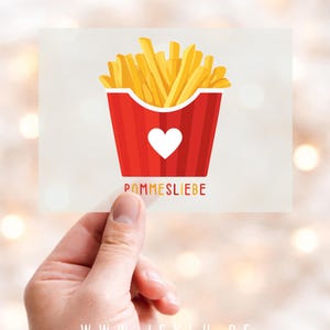 Könnte beinhalten: Eine rechteckige Karte mit einem roten Behälter mit goldenen Pommes Frites und einem weißen Herzen. Auf der Karte steht der deutsche Text "POMMESLIEBE". Die Karte wird in einer Hand vor einem verschwommenen Hintergrund gehalten.