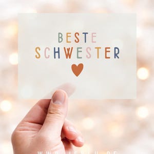 Könnte beinhalten: Eine rechteckige Karte, die in einer Hand gehalten wird, mit den Worten "BESTE SCHWESTER" in bunten Buchstaben und einem kleinen orangefarbenen Herz. Der Hintergrund ist mit Bokeh-Lichtern verschwommen.