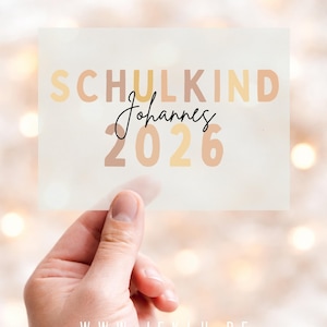 Könnte beinhalten: Eine transparente rechteckige Karte, die von einer Hand gehalten wird, mit den Worten "SCHULKIND Johannes 2026" in verschiedenen Braun- und Gelbtönen. Die Website-Adresse "WWW.JEKLU.DE" befindet sich unten.