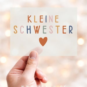 Könnte beinhalten: Eine Hand hält eine Karte mit dem deutschen Text "KLEINE SCHWESTER" in bunten Buchstaben, mit einem Herzsymbol. Die Website-Adresse "WWW.JEKLU.DE" befindet sich unten. Der Hintergrund ist mit Bokeh-Lichtern verschwommen.