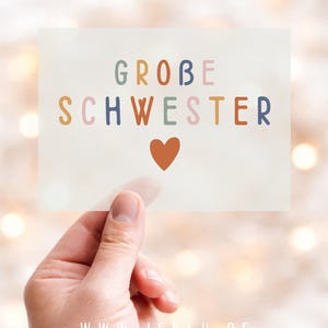Könnte beinhalten: Eine rechteckige Karte, die von einer Hand gehalten wird, mit den Worten "GROBE SCHWESTER" in bunten Buchstaben und einem kleinen orangefarbenen Herz darunter. Die Website-Adresse "WWW.JEKLU.DE" befindet sich unten.