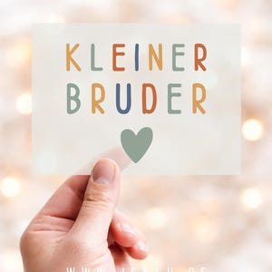 Könnte beinhalten: Eine rechteckige Karte mit dem Text KLEINER BRUDER in mehrfarbigen serifenlosen Buchstaben und einem salbeigrünen Herzsymbol unter dem Text. Die Karte wird vor einem unscharfen, hellen Hintergrund mit sanfter Bokeh-Beleuchtung gehalten.