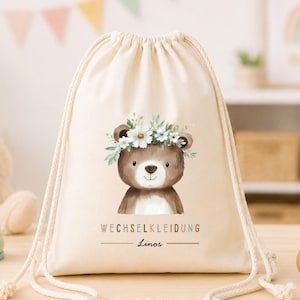 Könnte beinhalten: Ein cremefarbener Turnbeutel mit Kordelzug, verziert mit einer Illustration eines braunen Bären mit Blumenkranz. Unter dem Bären steht der Text WECHSELKLEIDUNG und der Name Linos. Der Beutel hat dicke, cremeweiße Kordeln und ein schlichtes Design.