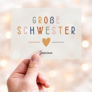 Könnte beinhalten: Eine rechteckige Karte mit dem Text GROBE SCHWESTER in bunten serifenlosen Buchstaben, einem senfgelben Herzsymbol und dem Namen Janina in schwarzer Schreibschrift auf einem hellen, halbtransparenten Hintergrund.