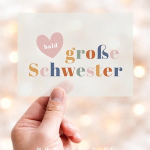 Könnte beinhalten: Eine rechteckige Karte, die von einer Hand gehalten wird, mit den Worten "bald große Schwester" in bunten Buchstaben. Ein rosa Herz mit dem Wort "bald" befindet sich über dem Text. Der Hintergrund ist ein weiches, verschwommenes Bokeh aus Lichtern.