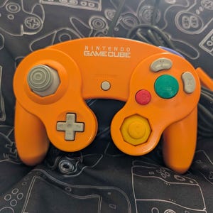 May include: A bright orange Nintendo GameCube video game controller featuring a gray analog stick, a gray D-pad, a large green A button, a red B button, gray X and Y buttons, a yellow C-stick, and a central gray Start/Pause button with a black cord.