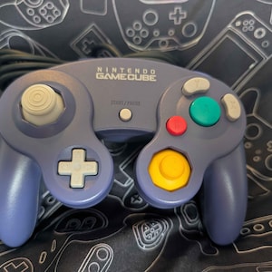 May include: A purple Nintendo GameCube video game controller featuring a white analog stick, a white D-pad, a large green A button, a red B button, a yellow C-stick, and gray X and Y buttons. The controller includes a Start/Pause button.