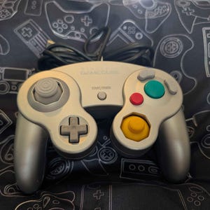 May include: A silver GameCube video game controller featuring a gray analog stick, a gray D-pad, a yellow C-stick, a green A button, a red B button, and a small gray Start/Pause button. The controller includes a black wired connection cable.