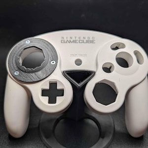 May include: A white Nintendo GameCube controller with a black analog stick housing and a black stand. The controller has a directional pad, buttons, and the text "NINTENDO GAMECUBE".