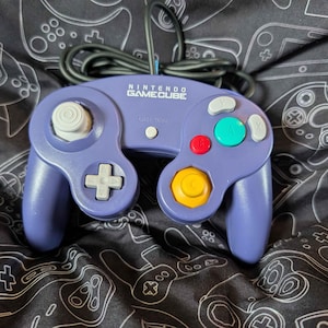 May include: A purple Nintendo GameCube video game controller featuring a white analog stick, a white D-pad, a yellow C-stick, a red B button, a green A button, and gray X and Y buttons. The controller includes a black wired cable extending from the top.