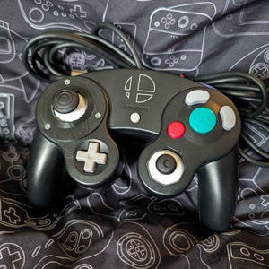 May include: A black wired game controller featuring a white Super Smash Bros. logo in the center. The device includes a large green A button, a red B button, two gray analog sticks, a gray directional pad, and additional gray action buttons.