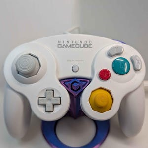 May include: White Nintendo GameCube controller with gray, red, teal, and yellow buttons. The controller has the words "NINTENDO GAMECUBE" printed on it. A purple and blue holder is attached to the bottom of the controller.
