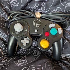 May include: A black wired video game controller featuring a Super Smash Bros logo in the center. The controller includes a gray analog stick, a gray D-pad, a large green A button, a red B button, a yellow C-stick, and gray X and Y buttons.