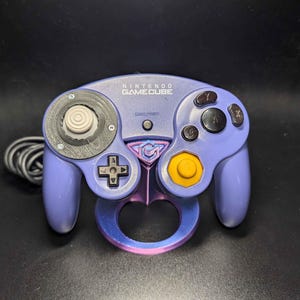May include: A purple Nintendo GameCube controller with a black cable. The controller has a circular analog stick, a directional pad, and several buttons. The controller has a custom-designed ring attachment in shades of purple and pink.