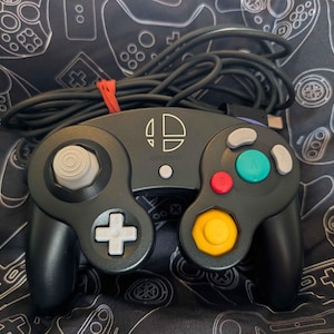 May include: A black video game controller featuring a white Super Smash Bros logo at the top center. The device includes a white analog stick, a white directional pad, a large green A button, a red B button, and a yellow C-stick with a long black cord.