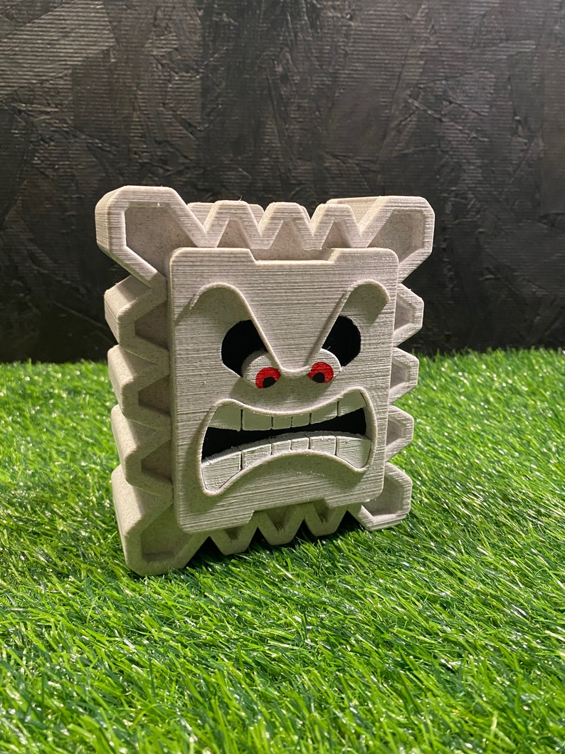 May include: A gray, 3D-printed Thwomp from the Super Mario video game series. The Thwomp has an angry expression with red eyes and a wide open mouth. The figure is sitting on a patch of green artificial grass.