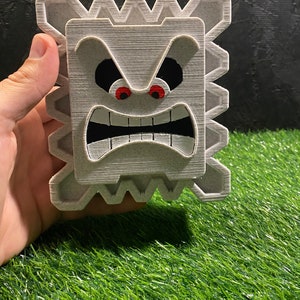 May include: A gray, 3D-printed Thwomp from the Super Mario video game series. The Thwomp has an angry expression with red eyes, a wide open mouth, and a textured surface. The Thwomp is held in a hand against a black background and green grass.