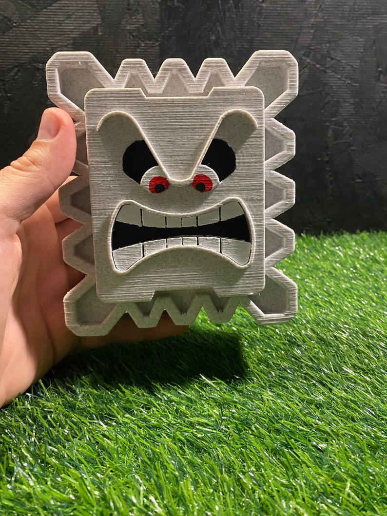 May include: A gray, 3D-printed Thwomp from the Super Mario video game series. The Thwomp has an angry expression with red eyes, a wide open mouth, and a textured surface. The Thwomp is held in a hand against a black background and green grass.