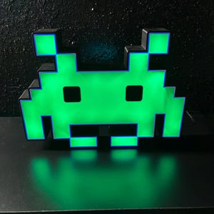 Puede incluir: Una lámpara verde pixelada con forma de alienígena y borde negro. La luz emite un brillo verde brillante, con un contorno azul. La lámpara está sobre una superficie negra y el fondo es oscuro.