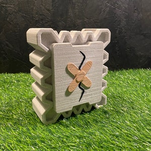 May include: A gray, 3D-printed decorative object with a square face featuring a brown bandage and a black crack. The object has a textured, layered design and sits on a green surface. The overall shape is a cube.