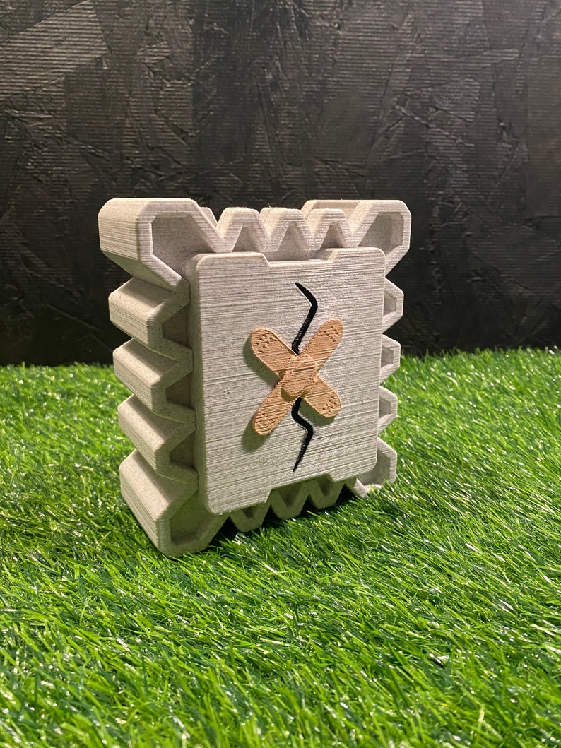 May include: A gray, 3D-printed decorative object with a square face featuring a brown bandage and a black crack. The object has a textured, layered design and sits on a green surface. The overall shape is a cube.