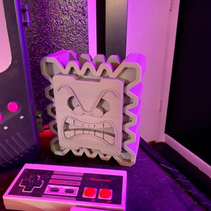May include: A light gray Thwomp statue with an angry face, next to two Nintendo controllers. The controllers are black and white with red buttons and the Nintendo logo. A black and gray console is also visible.