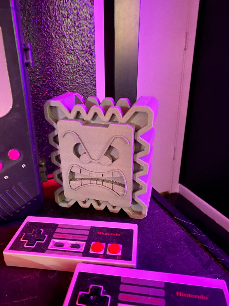 May include: A light gray Thwomp statue with an angry face, next to two Nintendo controllers. The controllers are black and white with red buttons and the Nintendo logo. A black and gray console is also visible.