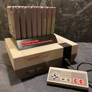 May include: A vintage Nintendo Entertainment System console with a controller and a game cartridge holder. The console is beige and gray, with red lettering. The controller is gray with red buttons. A collection of game cartridges are in the holder.