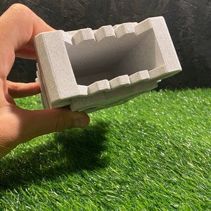 May include: A light gray rectangular ashtray with a textured surface. The ashtray has a series of notches along the top edge and is held against a green and black background.