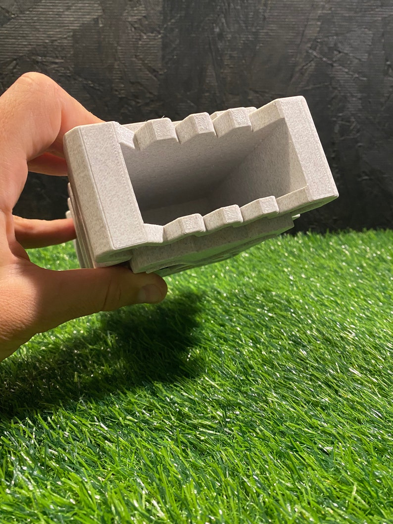 May include: A light gray rectangular ashtray with a textured surface. The ashtray has a series of notches along the top edge and is held against a green and black background.