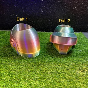 Puede incluir: Dos objetos iridiscentes con forma de casco, impresos en 3D, etiquetados como "Daft 1" y "Daft 2", están sobre una superficie verde. Los cascos tienen un brillo metálico arcoíris, con colores que cambian de rosa a verde y amarillo. El fondo es oscuro.