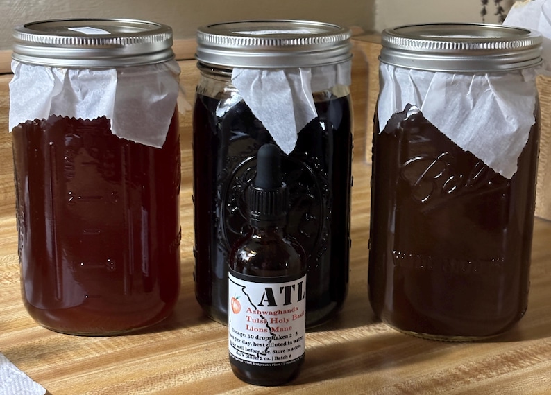 May include: Three glass jars filled with dark liquids, covered with paper and metal lids. A small bottle with a dropper and a label that reads "ATL" is in front of the jars. The label also lists ingredients and instructions.