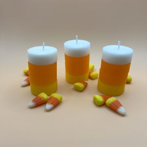 Può includere: Tre candele a strisce arancioni e gialle con cime bianche, circondate da mais dolce.