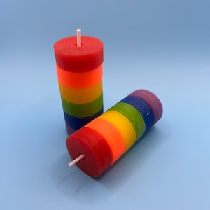 Puede incluir: Dos velas de capas de arcoíris con una mecha blanca. Las velas están apiladas en un patrón de arcoíris con capas rojas, naranjas, amarillas, verdes, azules y moradas.