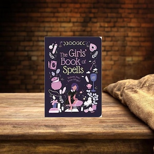 May include: A hardcover book titled "The Girls' Book of Spells" with a dark purple cover. The cover features illustrations of magical items and a girl casting a spell. The book is on a wooden surface with a brick background.