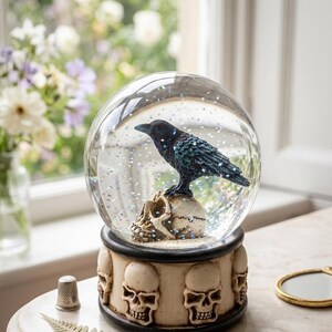 May include: A clear glass snow globe containing a black raven perched on a human skull, filled with iridescent glitter. The base is a cylindrical cream-colored stand decorated with a repeating pattern of sculpted human skulls and dark trim.