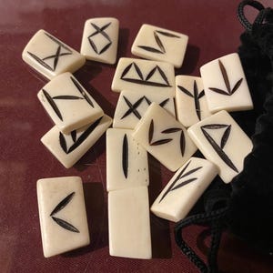 May include: A set of white bone runes with black markings, in a black velvet bag. The runes are arranged in a pile, with some of them overlapping.