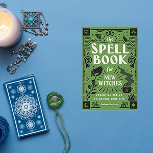 May include: A still-life composition on a blue surface features a lit candle, a deck of tarot cards, and a green-covered spell book. The book, titled "The Spell Book for New Witches," is surrounded by various mystical accessories, including jewelry and a small glass bottle.