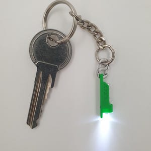 May include: A silver key attached to a key ring and chain, with a small green LED flashlight. The flashlight is rectangular and has the word "LIXY" printed on it. The light is on and shining brightly.