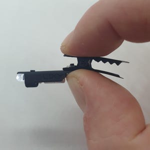 May include: A black clip-on LED light with a white LED bulb. The light is held in a hand.