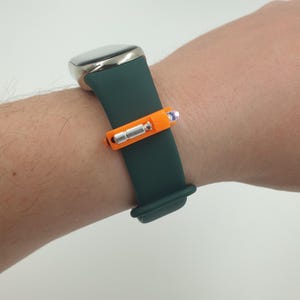 May include: A dark green watch strap with a silver watch face and an orange accessory. The accessory features a silver cylinder and a small, glowing blue light. The watch is on a wrist.