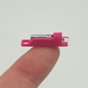 May include: A small, pink plastic component with the word "LIXY" printed on it. It has a silver metal piece and a clear, rounded end. The component is held between a finger and thumb against a plain white background.