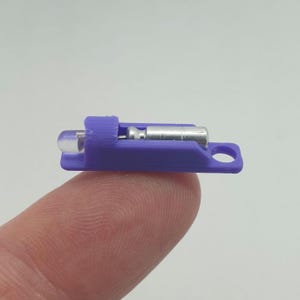 Puede incluir: Un pequeño pestillo de perno morado impreso en 3D con un componente metálico plateado y un extremo redondeado transparente. El pestillo se sostiene en un dedo, mostrando su tamaño y diseño en miniatura. El pestillo tiene un pequeño agujero en el extremo.