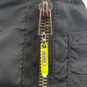 Op de afbeelding: Close-up van een zwarte jasrits met een messingkleurige metalen rits en een neongele ritstrekker. De rits is gedeeltelijk gesloten en toont de tanden en het ontwerp van de ritstrekker.