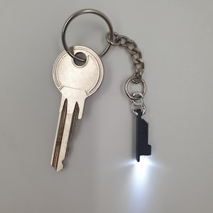 May include: A silver key and a black keychain with a small, rectangular LED light. The keychain is attached to a metal key ring and a short chain. The LED light is illuminated, emitting a bright white glow.